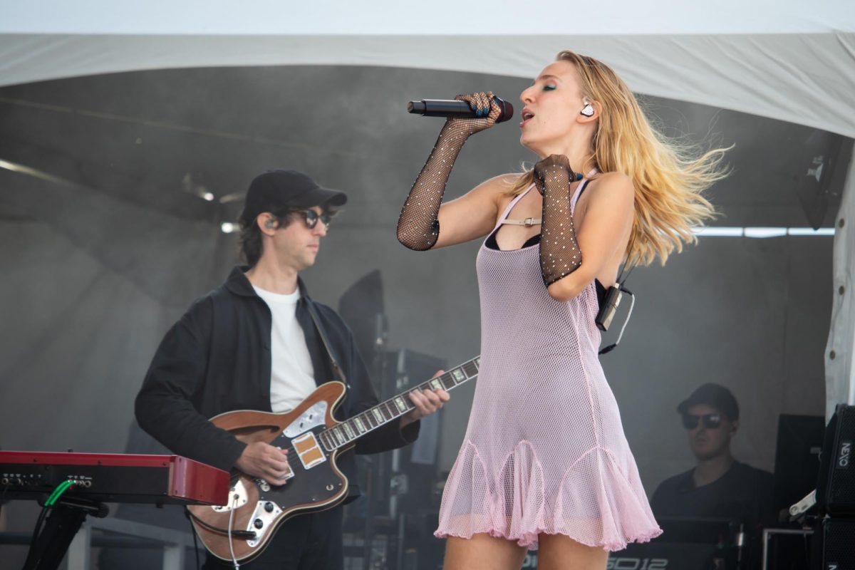A person onstage wearing a light pink dress sings into a microphone in front of a guitarist.