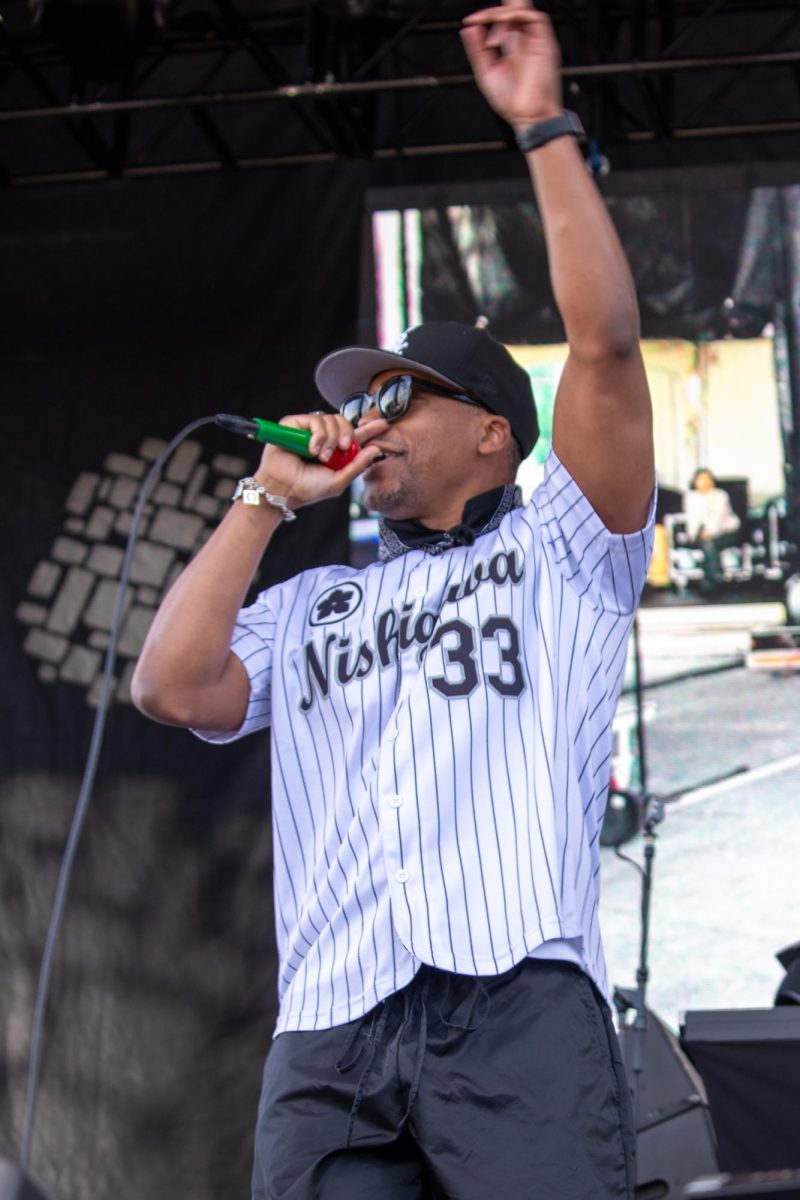 A person wearing a white striped baseball jersey and hat sings into a multi-colored microphone while raising their left arm upward.