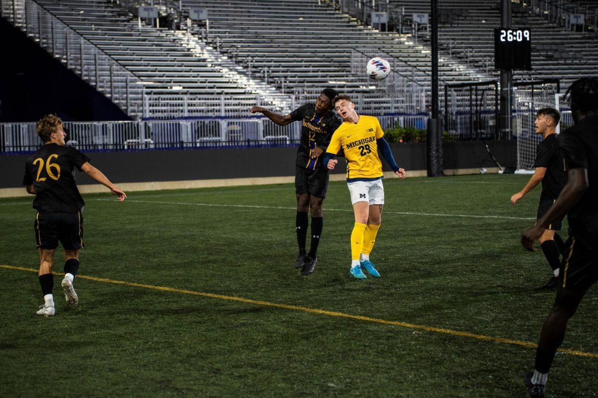 A Northwestern defender wearing black and Michigan forward wearing yellow and white jump in the air and collide heads.