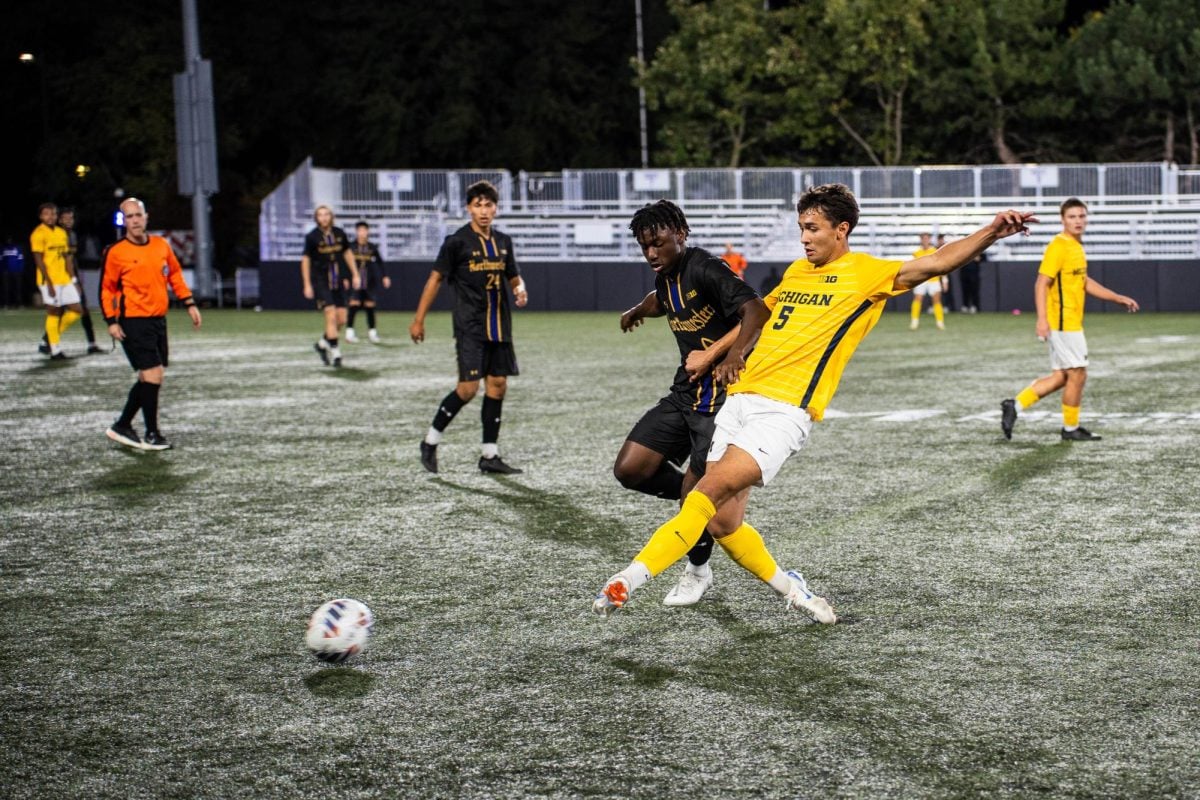 A Northwestern midfielder wearing black extends his arm in front of a Michigan opponent. The Michigan player’s leg is extended as he kicks the ball.