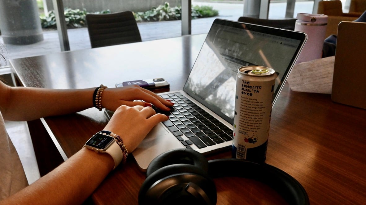 A student typing on a laptop with headphones and a drink beside the computer on a brown wooden table.