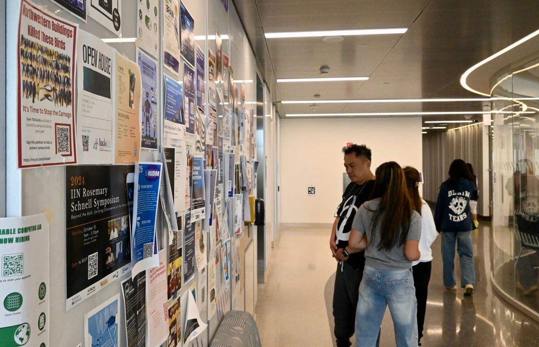 Two individuals look at flyers along a bulletin wall.