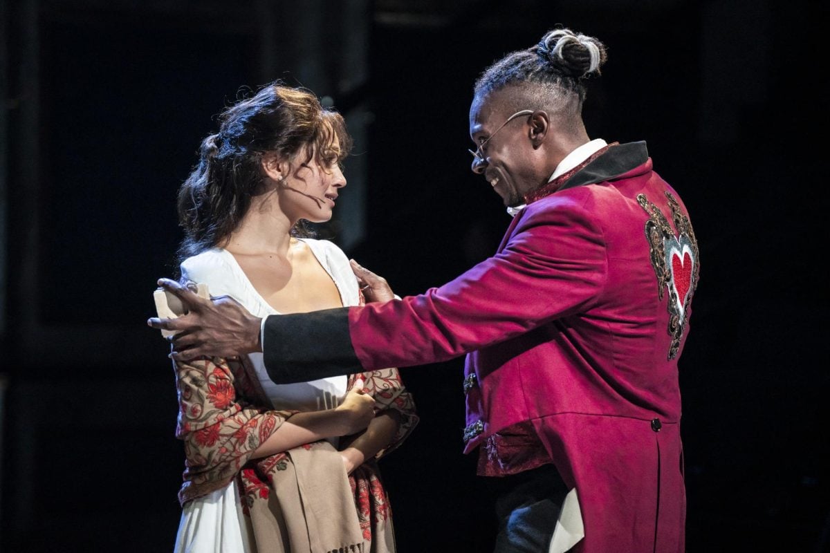 Female actor in a white dress being held by male actor wearing red jacket on stage.