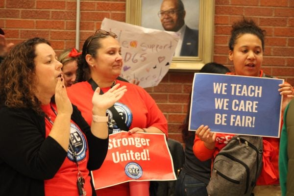 One woman in a red shirt claps, another holds a sign saying “Stronger United!” and another holds a sign saying “We teach, we care, be fair!”