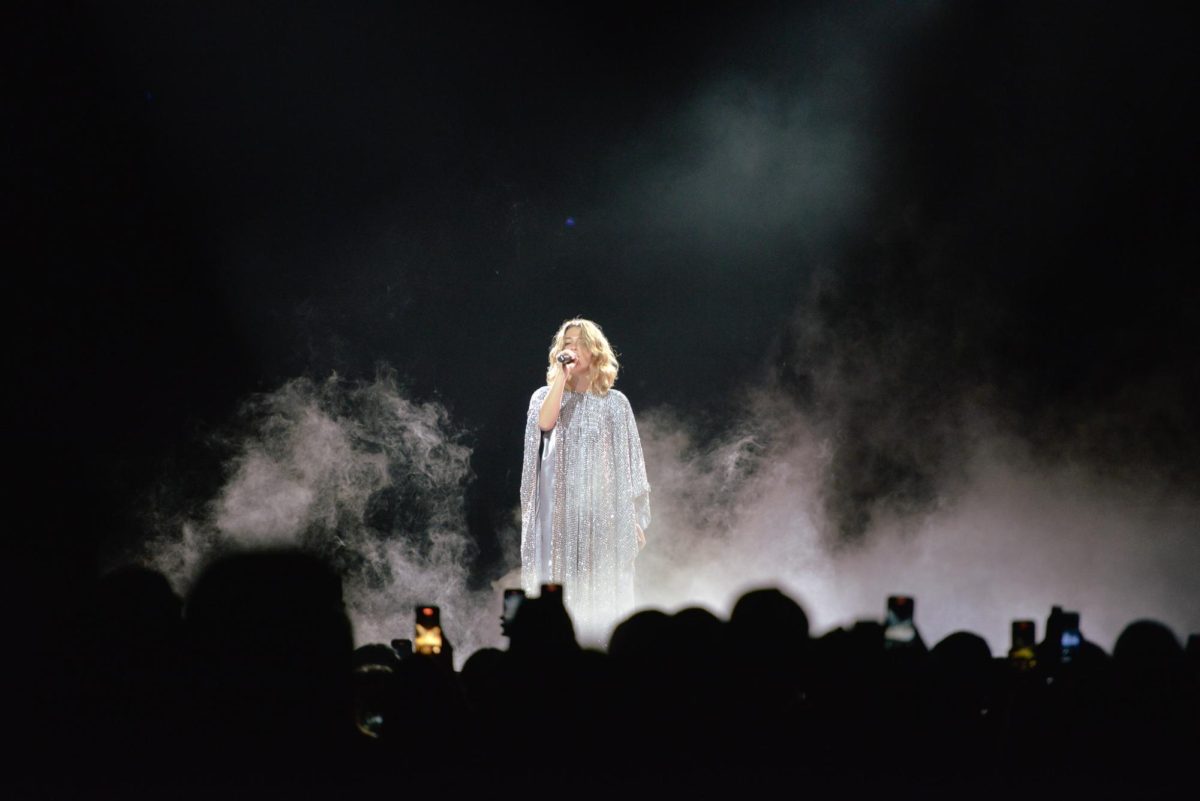 Rogers holds up a microphone in a floor-length silver robe surrounded by a cloud of fog.