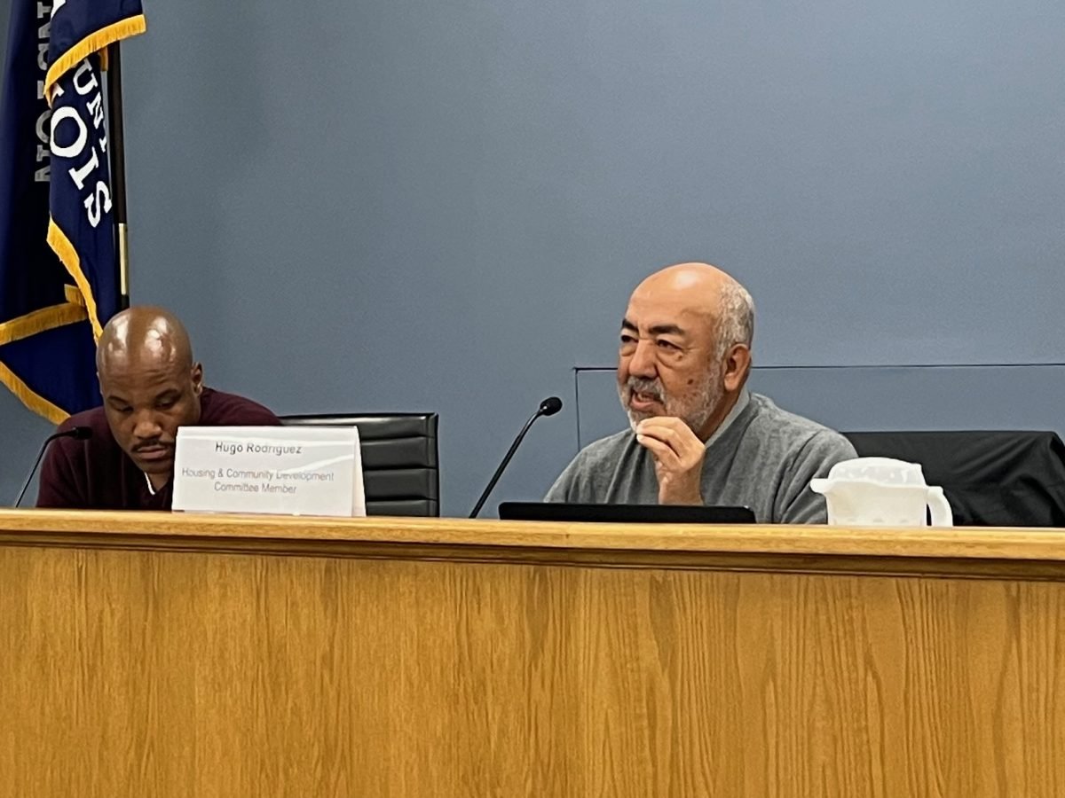 A man sitting behind a desk speaks while another man sits beside him.