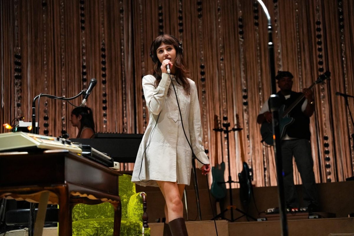 A singer wearing a cream-colored dress and headphones sings into a microphone.