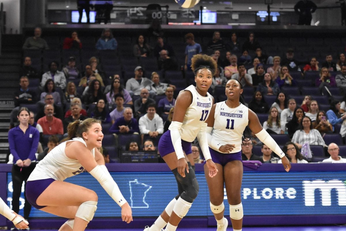 An athlete in a white jersey hits a volleyball as two teammates watch.