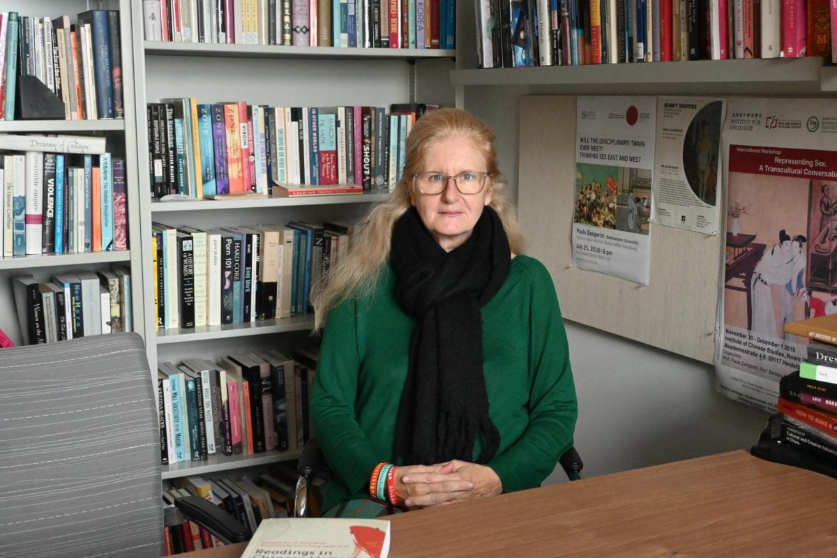 Woman wearing green sweater and black scarf sitting at her desk with bookshelves behind her