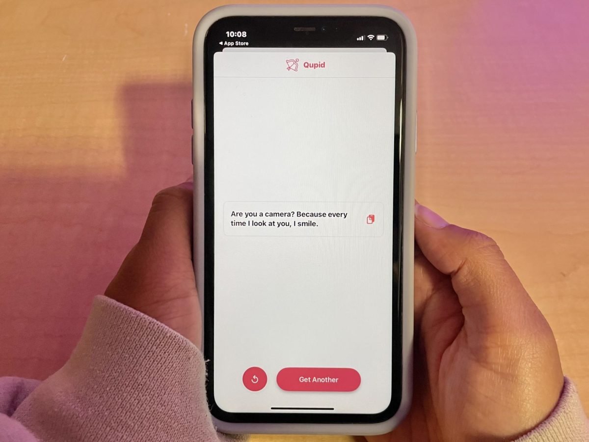 Two hands hold an iPhone opened to the app Qupid. There is a pickup line on screen that reads “Are you a camera, because everytime I look at you I smile.”
