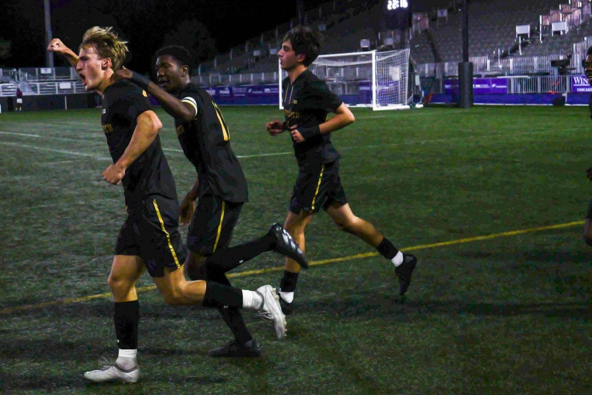 Three Northwestern players, all wearing black uniforms, run onto the field. The player running in the front pumps his fist, while the player behind him pats him on the back.