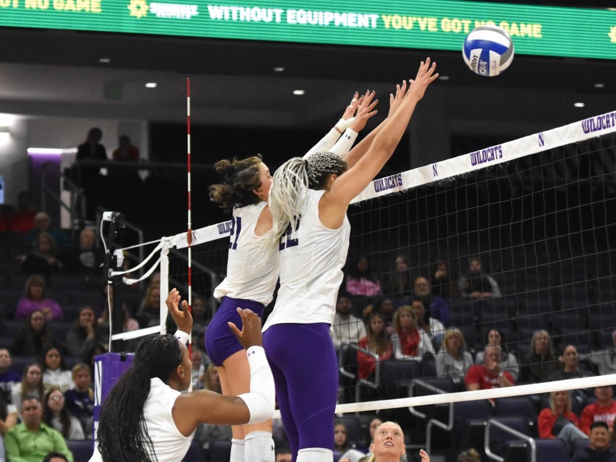 Two athletes in white jerseys block a volleyball at the net.