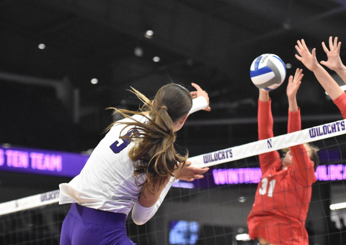 An athlete in a white jersey spikes a volleyball past two defenders.