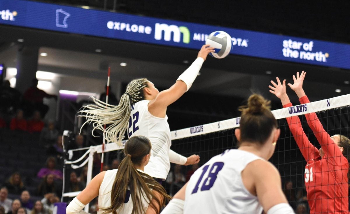 An athlete in a white jersey jumps to hit a volleyball over an opposing player.