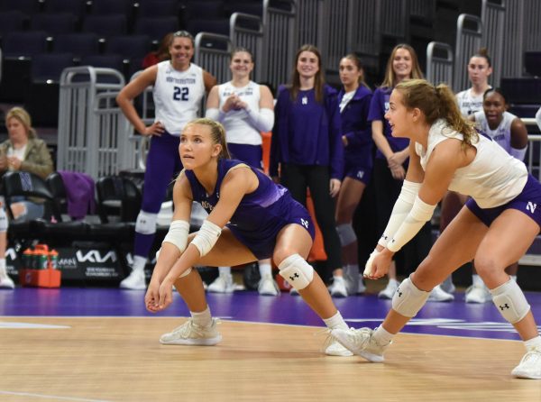 Two athletes in white and purple jerseys crouch and prepare to hit a volleyball.