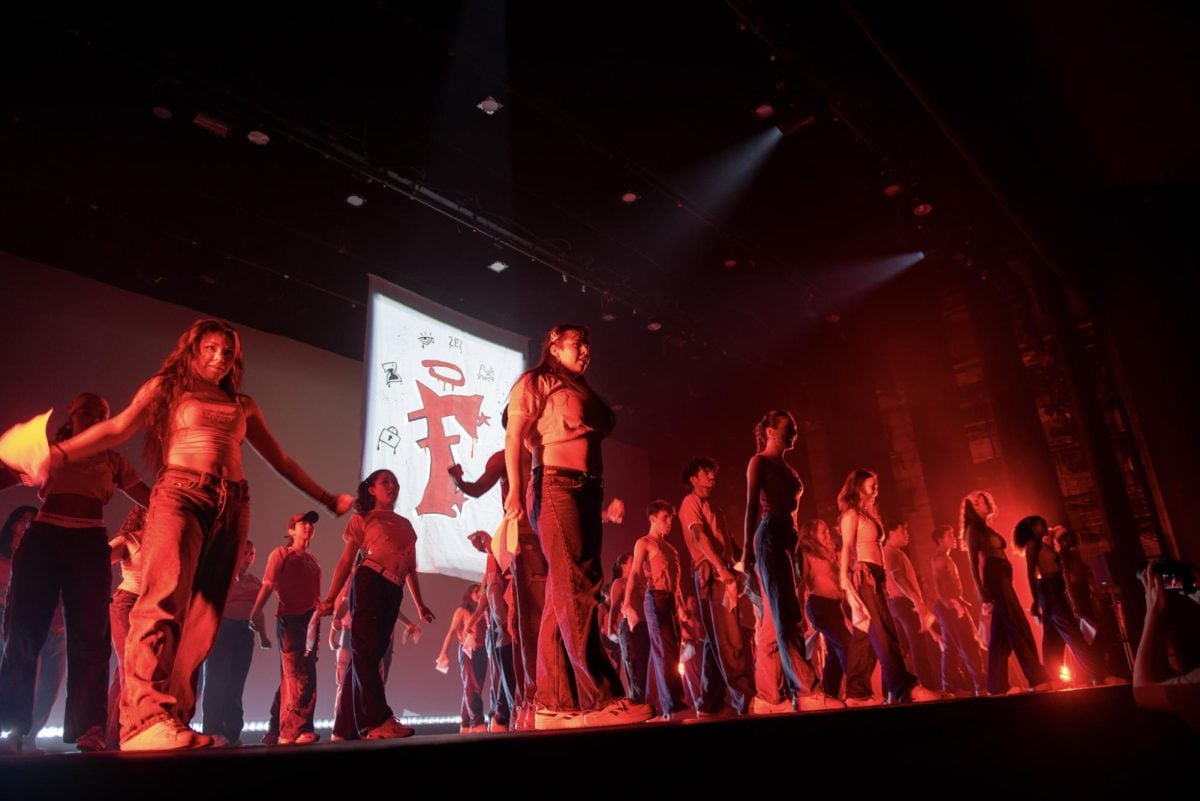 Dancers fill the red-lighted stage under a spotlighted banner.