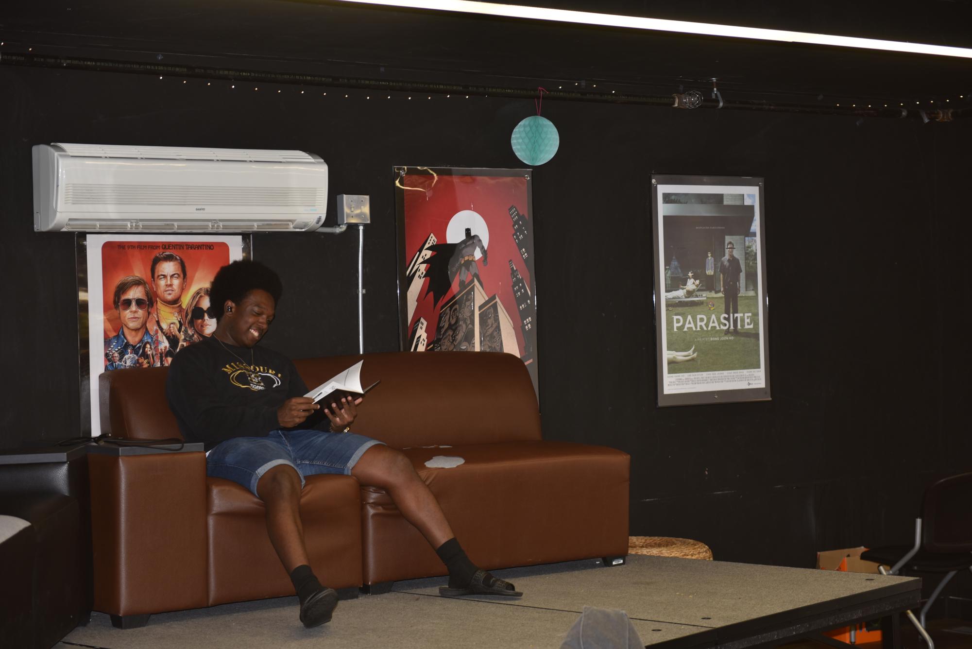 A student sits on a couch and smiles as he reads a book in East Fairchild. There are three posters on the wall behind him.