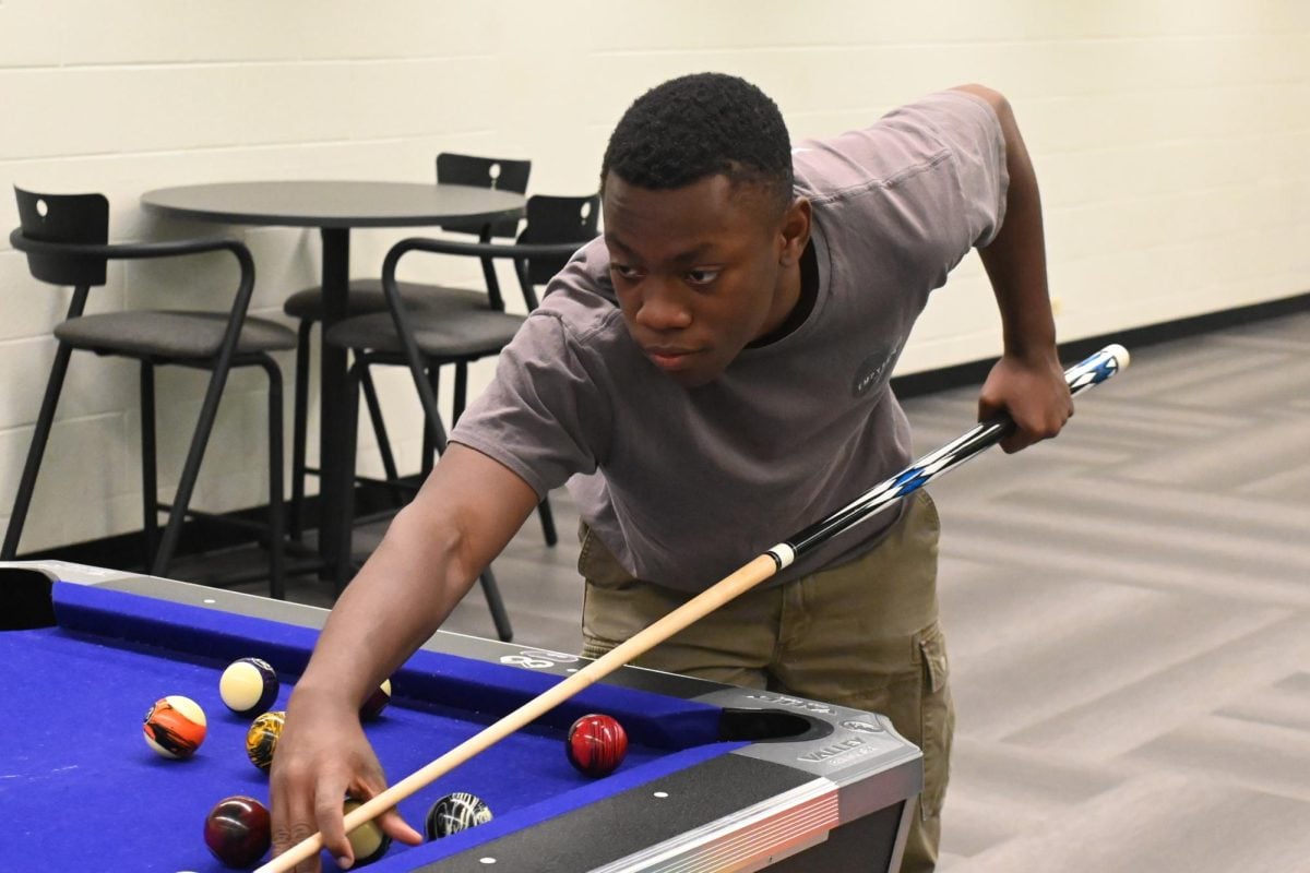 A student leans over a table with a cue stick in hand. His right finger is over the cue stick, and he is looking at the table.