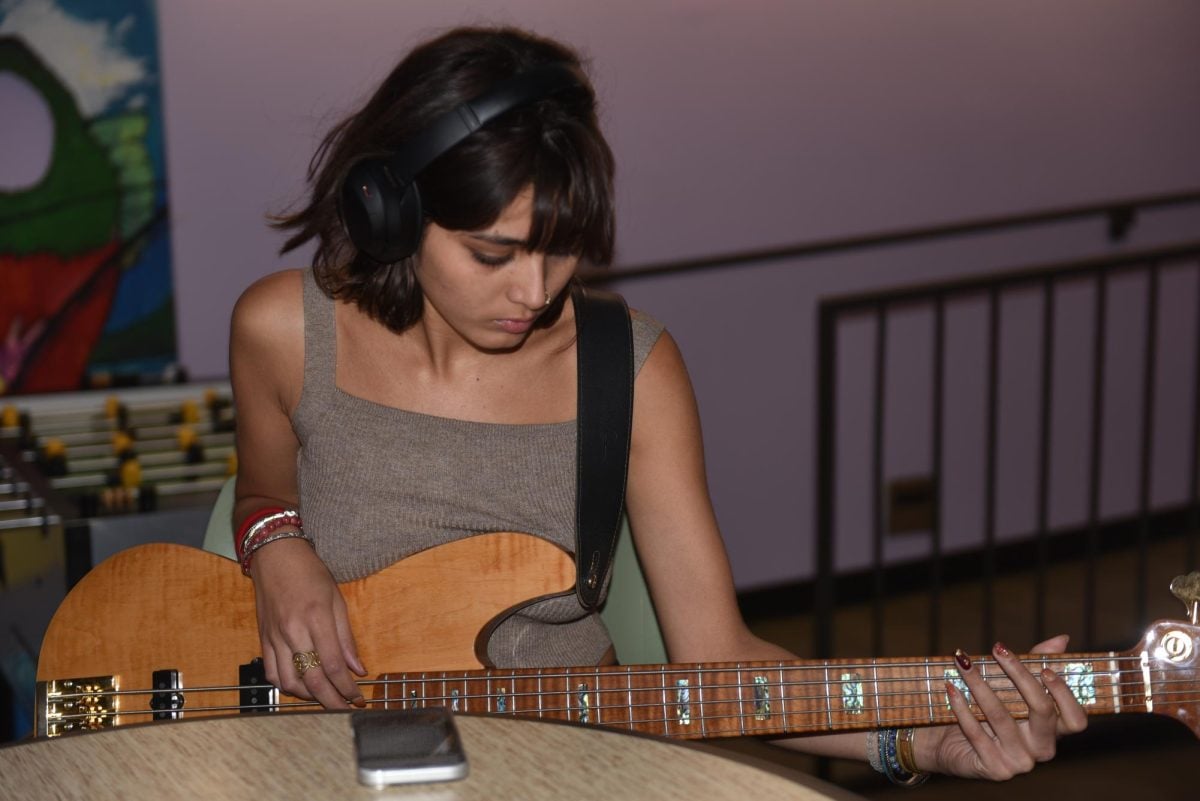 A girl who is sitting in a chair with her headphones on looks down as she plays her guitar. 