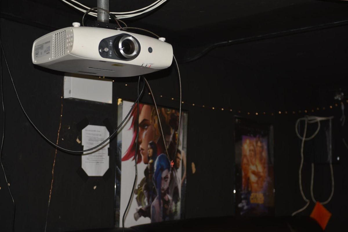 A projector is plugged into the wall, and in the background are two movie posters.