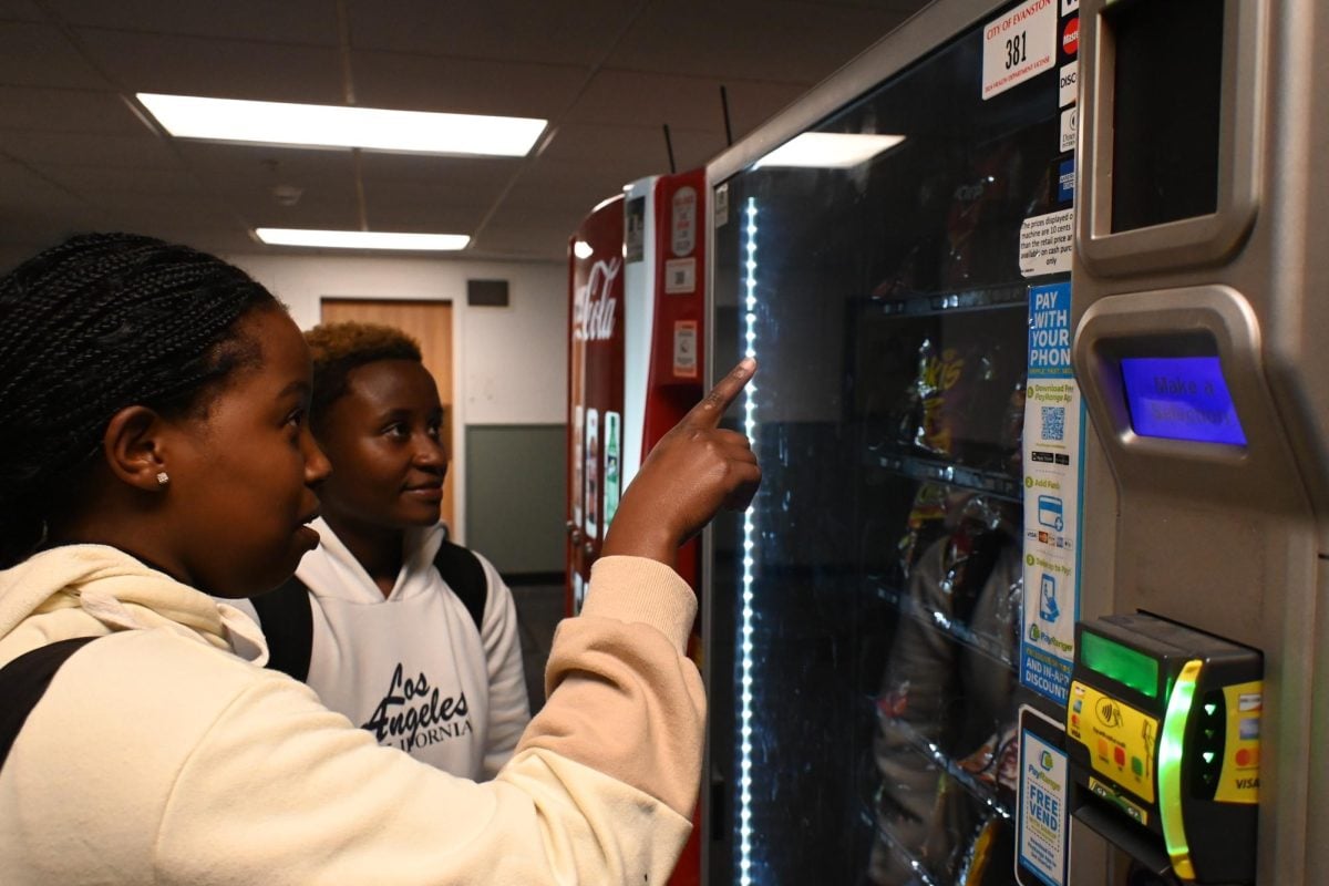 One student looks directly at a vending machine. The friend next to her is pointing at the vending machine while she is looking at it.