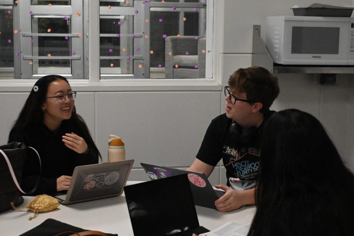 A student with glasses is sitting at a table on her computer and is smiling and laughing. The boy with glasses who sits next to her is speaking and staring directly at her.