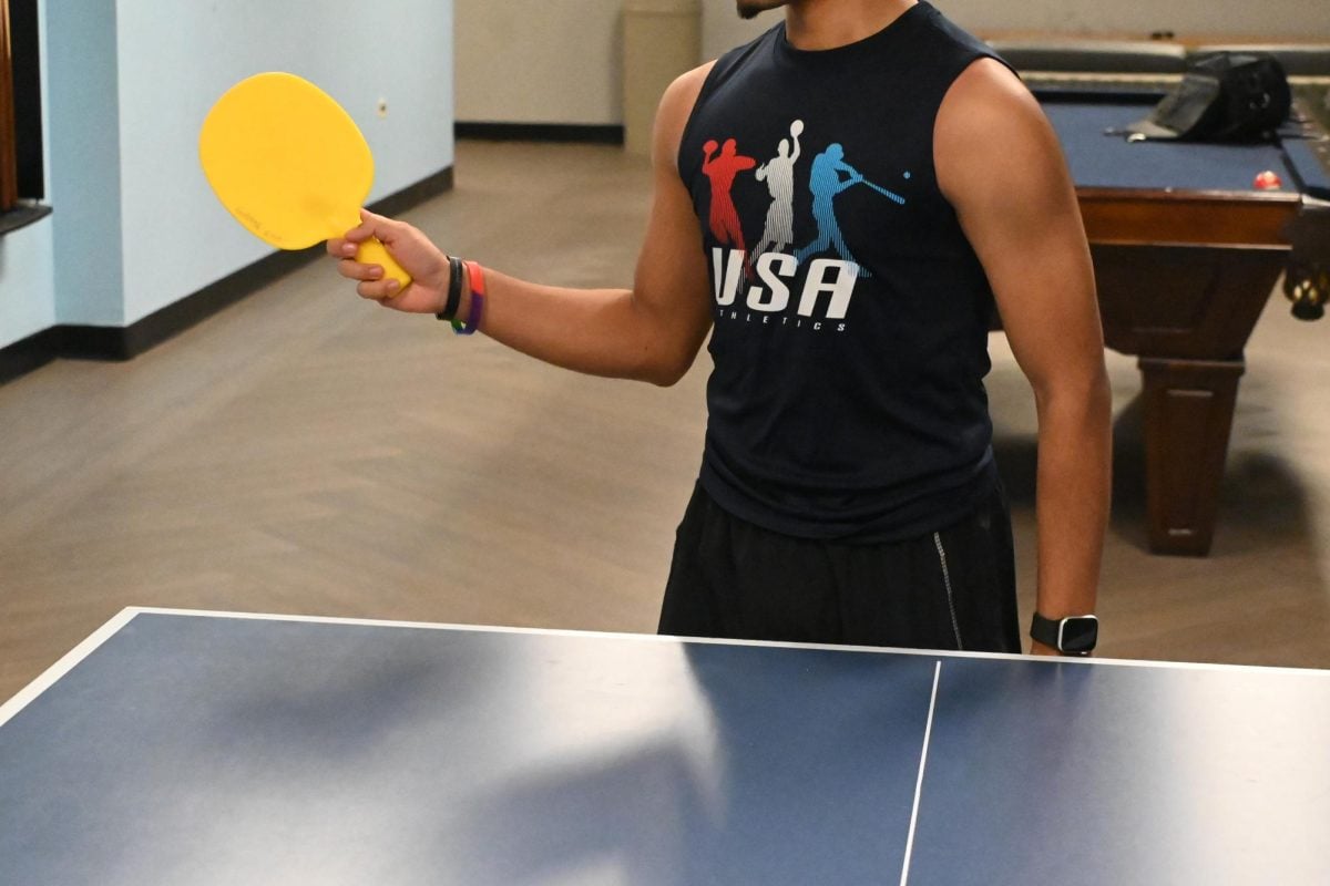 A student wearing a “USA Athletics” shirt is about to hit a ping-pong ball with the table tennis racket.