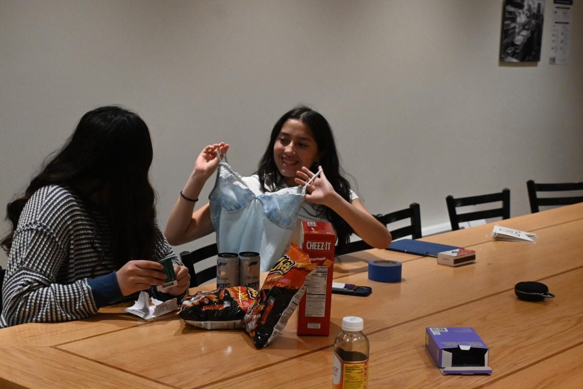 A girl is looking at her friend on her right who is holding up a Halloween bustier. They are both seated at a table with Cheetos, Celsius drinks, Cheez-It crackers, a phone, tape and other miscellaneous items.