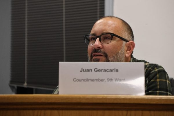 A man sits behind a name placard reading “Juan Geracaris, Councilmember, 9th Ward.”