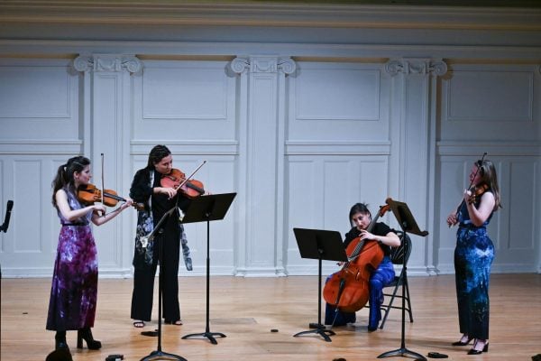 From left to right a woman stands playing violin, the next plays viola, the next sits while playing cello and the final woman also stands while playing violin.