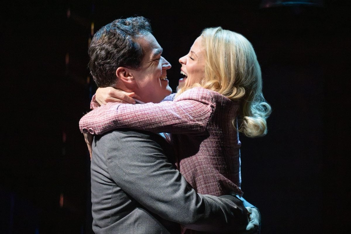 Man wearing a gray suit jacket and a blonde woman wearing a pink blazer hug and smile at each other.