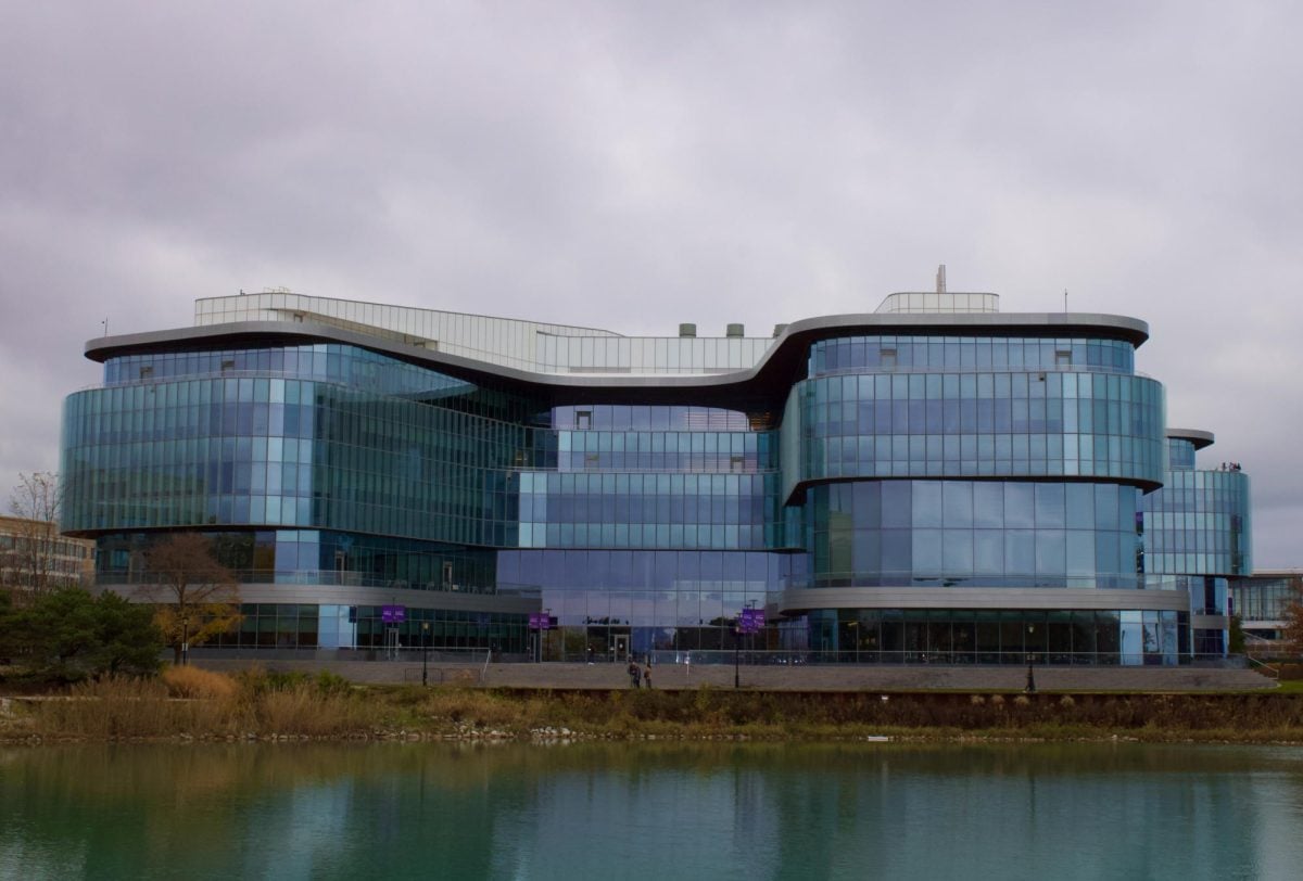 Long shot of Kellogg across the center of the Lakefill, semi-transparent glass windows reflecting gray sky with people passing by in front of back entrance steps. 