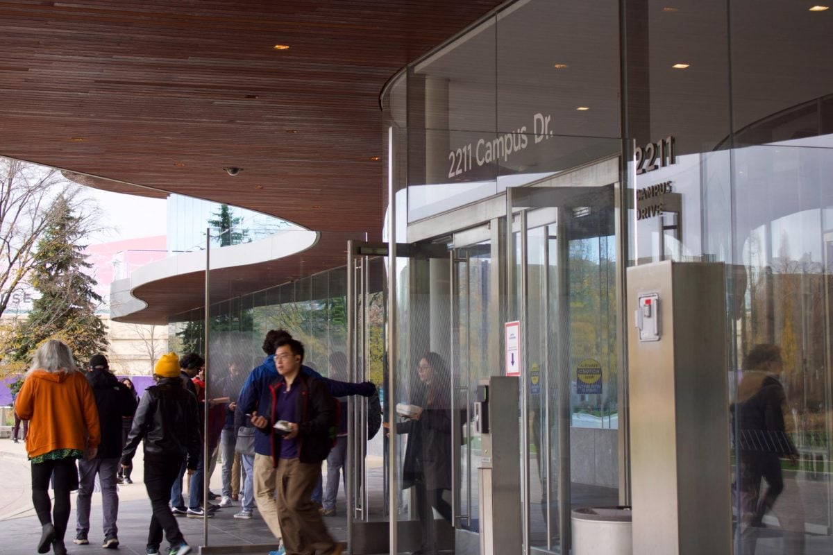 People walking out of Kellogg’s main entrance, with imprinted gray letters above glass doors indicating the building’s address, “2211 Campus Drive.”