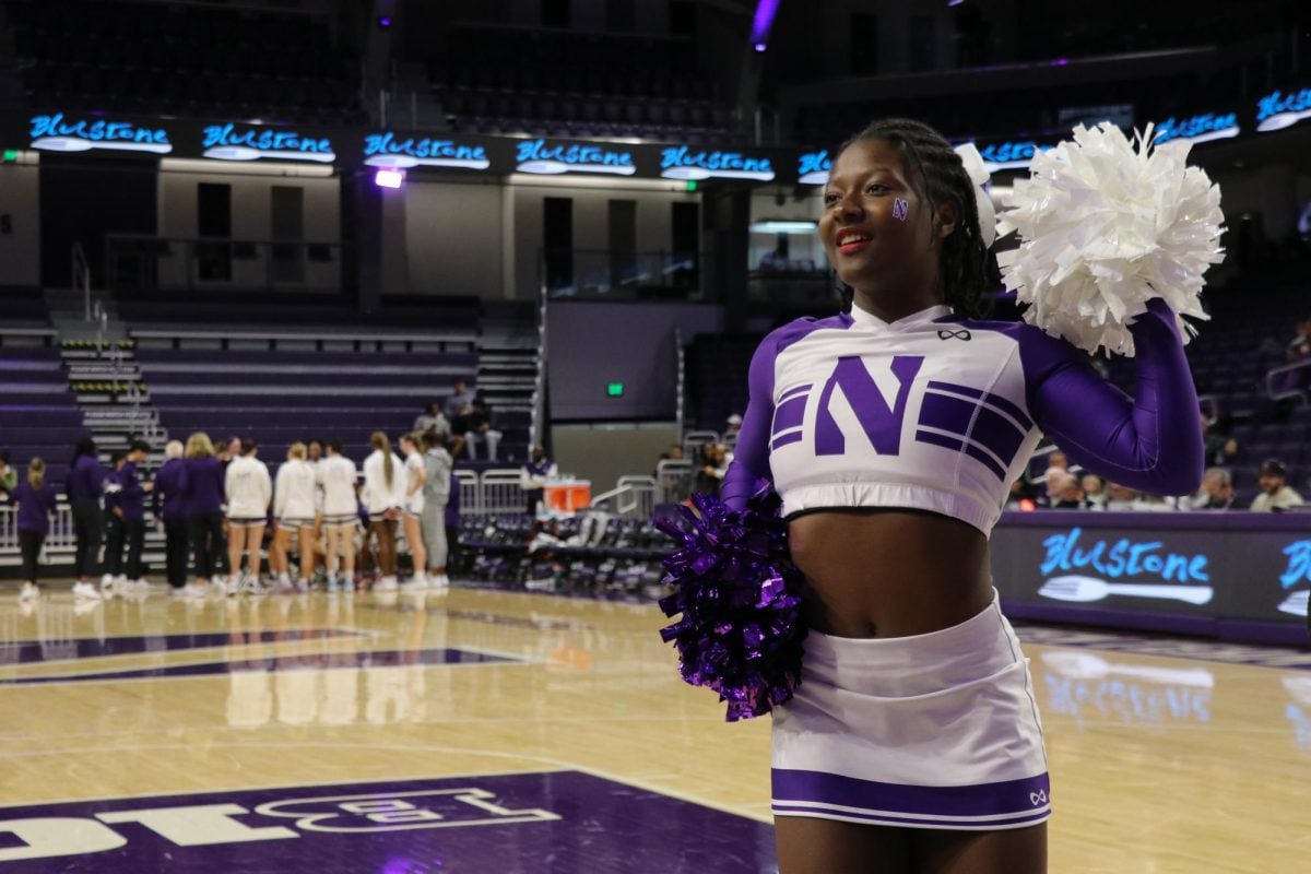 A cheerleader holds up white and purple pom poms in front of the team huddle.