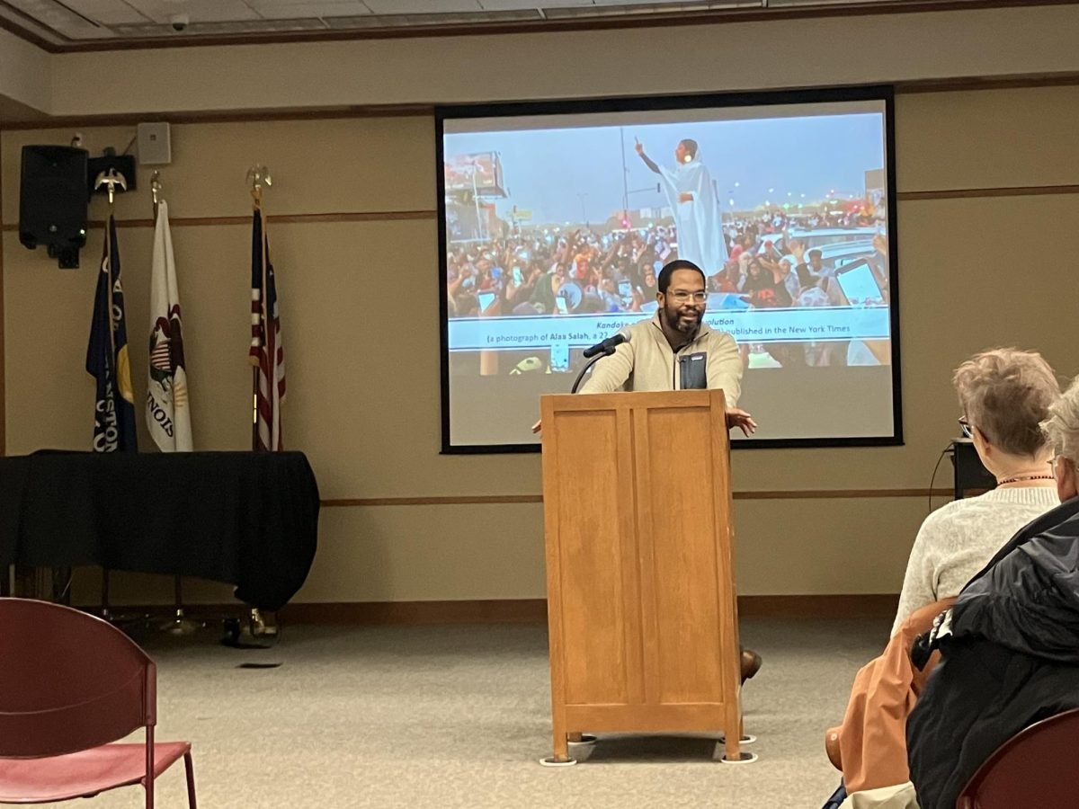 Alden Young, an associate professor of history and global affairs at Yale University stands in front of a podium with a presentation picturing protests in Sudan behind him.