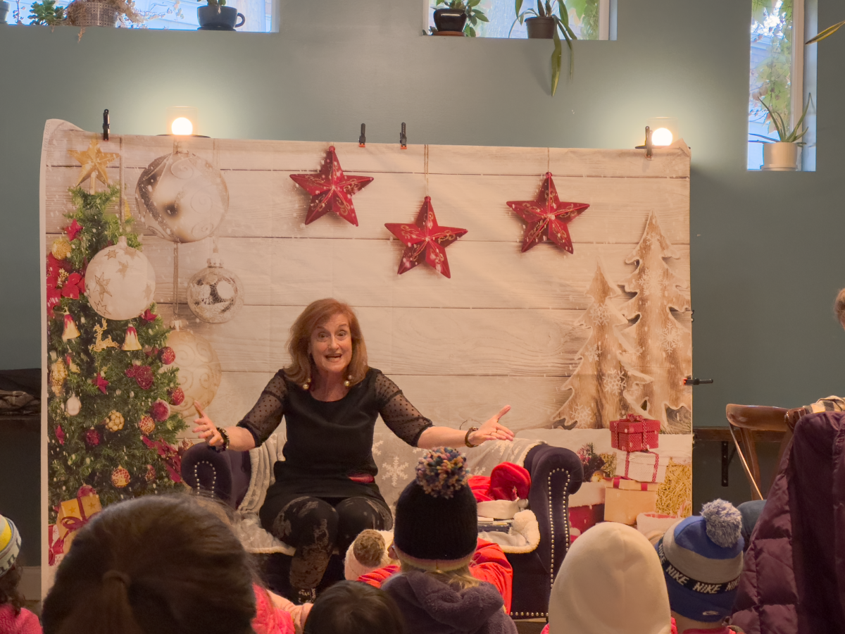 A woman sits on a couch in front of a holiday backdrop, speaking to a group of children.