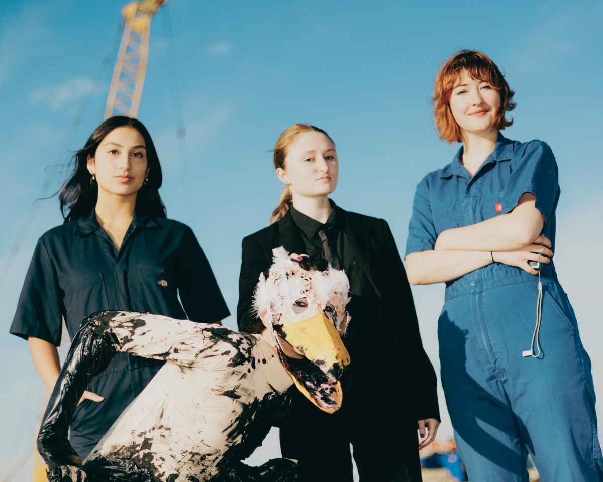 Three people and an anthropomorphic seagull stand in front of a blue sky and construction site.