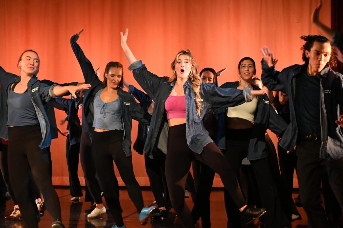 A large group of people perform a choreographed dance move on stage in front of an orange curtain.