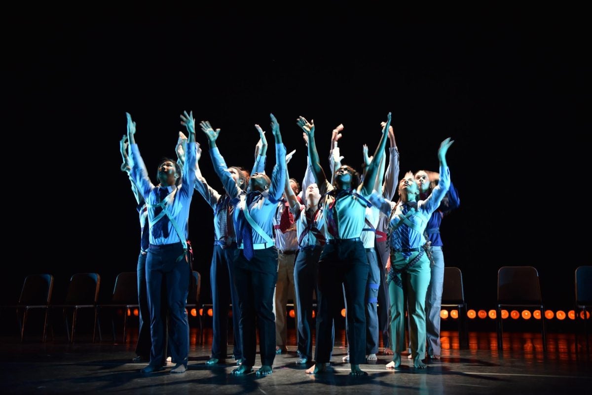 A group of people wearing uniforms — light blue collared shirts, navy pants, and red ties — clustered together, both hands stretched out to the sky.