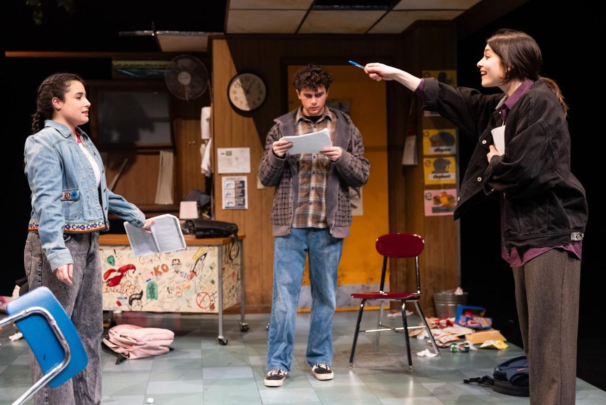 Three actors stand in a triangle onstage. The floor is littered with garbage and a backpack.