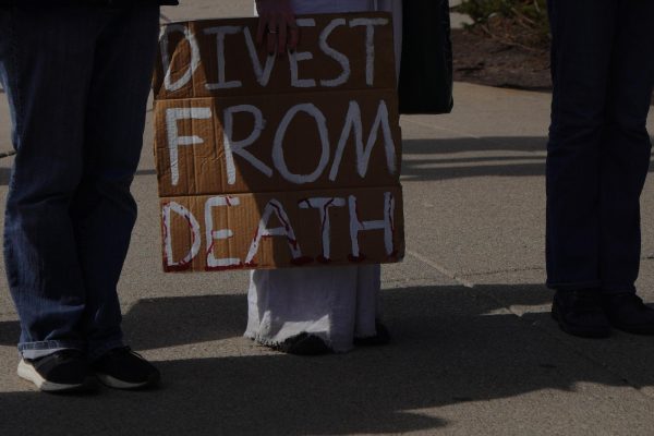 A demonstrator holds a cardboard sign reading &ldquo;Divest from death.&rdquo;