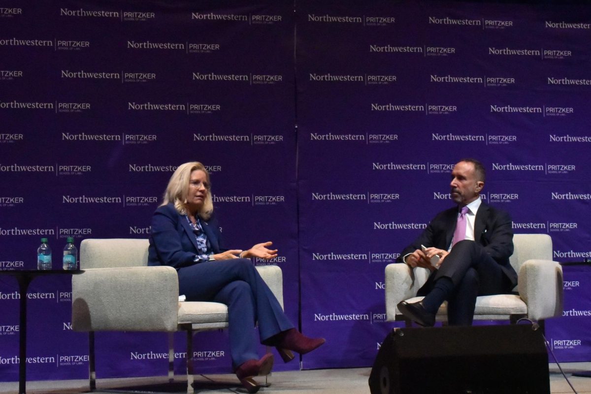 A woman and a man in suits sit in chairs in front of a purple background.