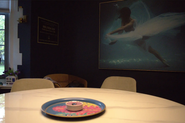 A room with a painting of an ascending dancer next to a plaque for Melissa Doi, both overlooking a table with colorful plates in the center.