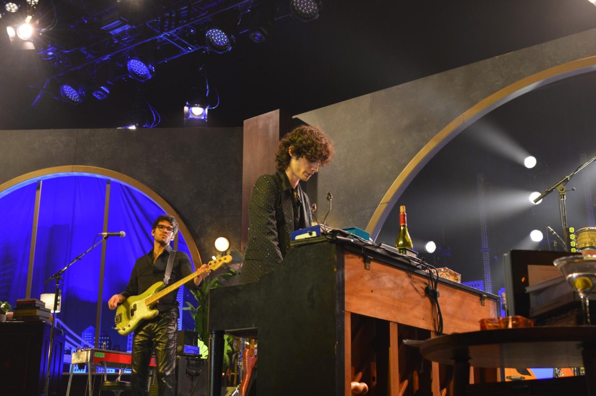 Man stands over piano with guitar player in the background. 