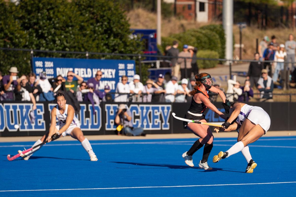 A player in a white jersey hits a field hockey ball as two other players watch. 
