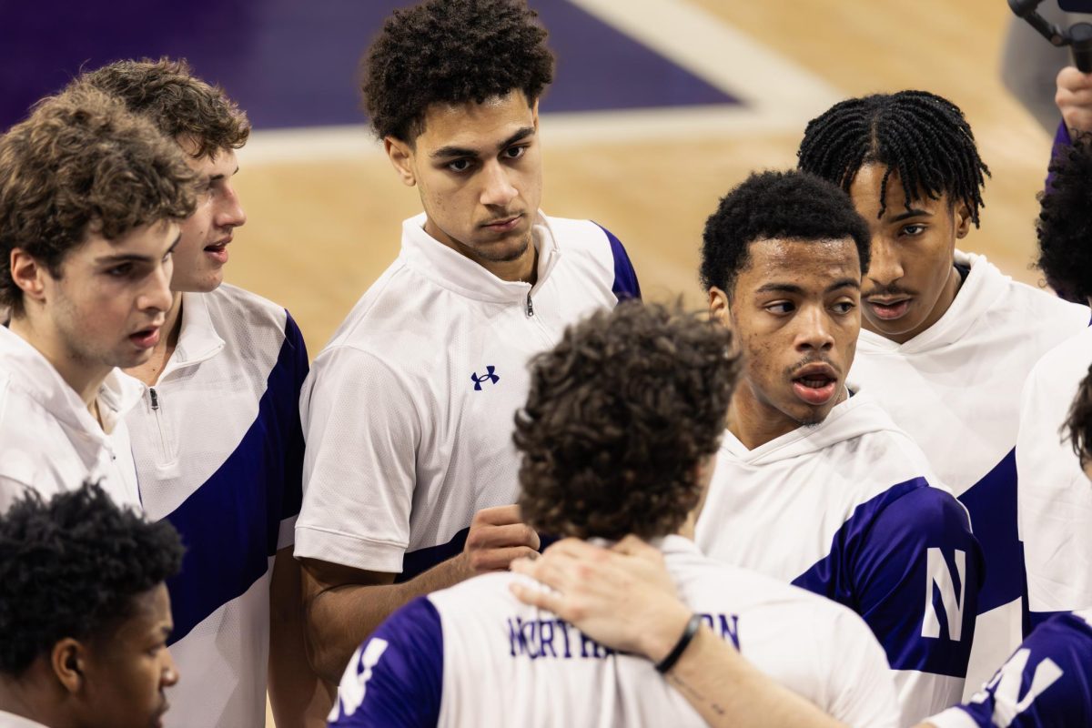 A group of Northwestern men&rsquo;s basketball players huddle together during warmups, listening intently and standing close with focused expressions. 