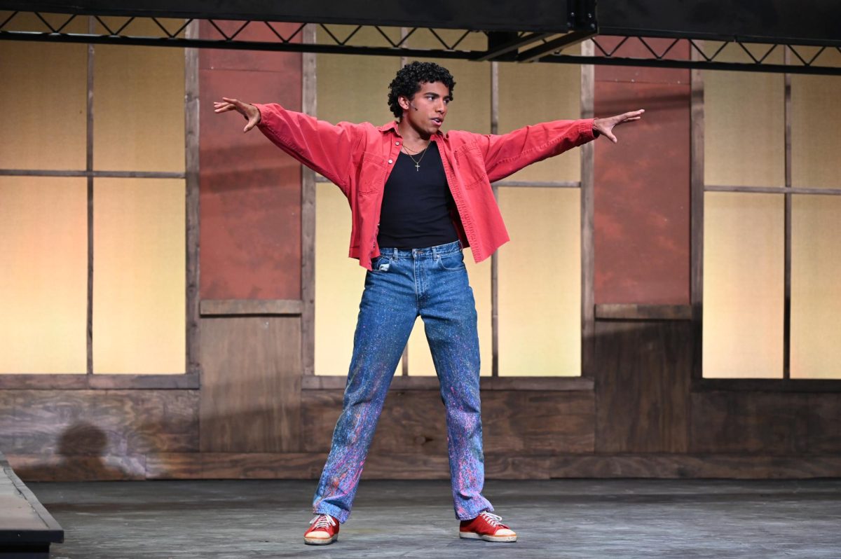 A man in a red shirt, black tank top, shimmery blue jeans and red sneakers performs on stage.
