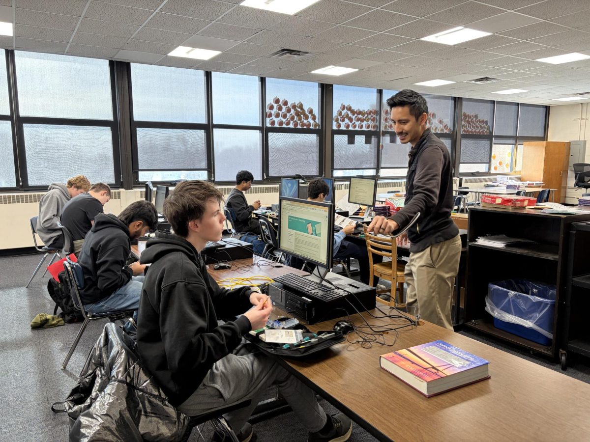 Students sit at desks with computers carefully assembling Ethernet cables with the guidance of their teacher.
