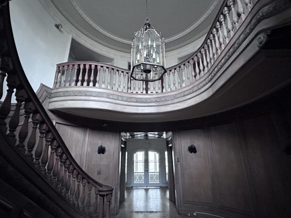 An entryway to a mansion includes a curved wooden staircase and a large chandelier.