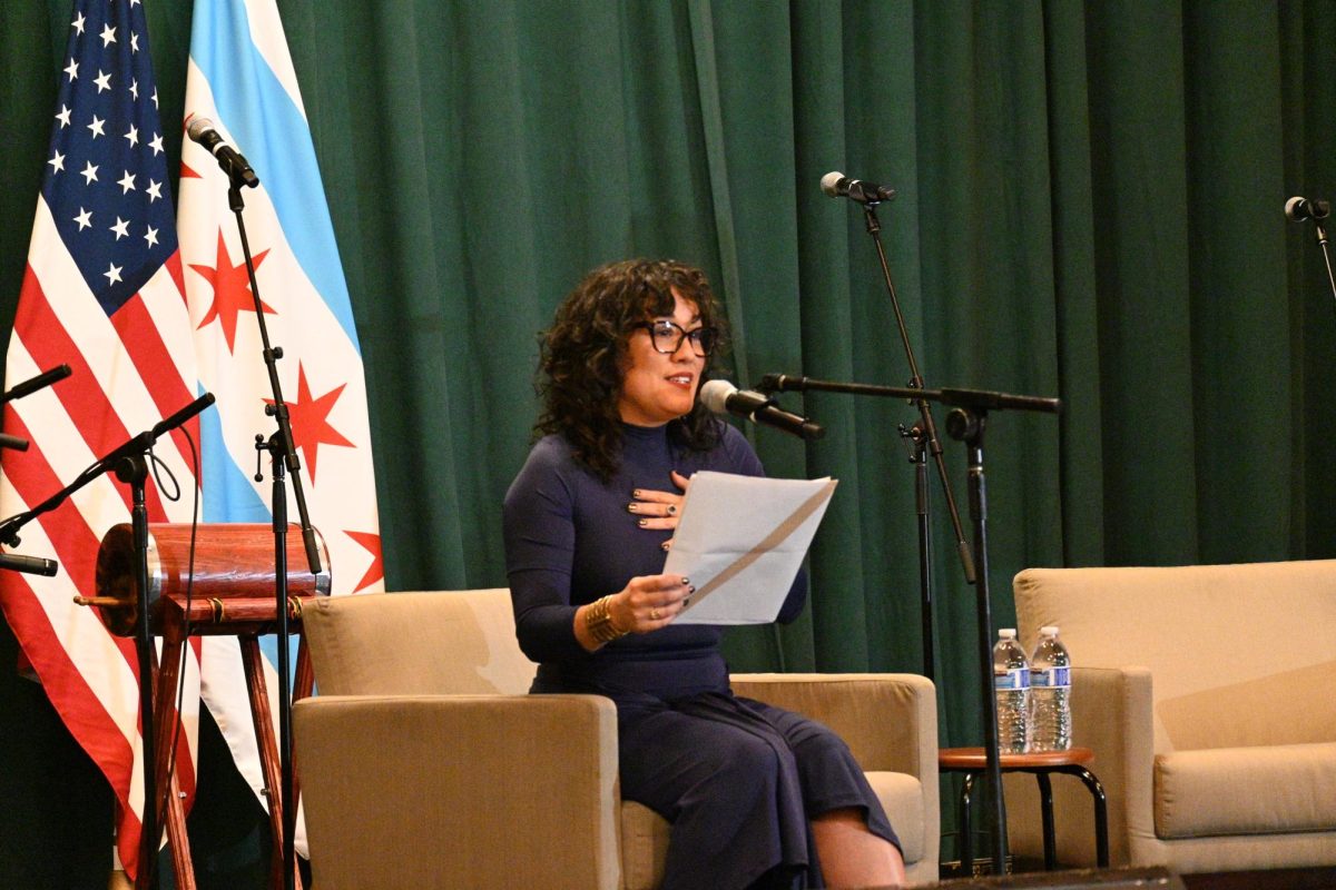 Person sits in a beige chair reading poetry into a microphone, flanked by an American flag and green curtain.